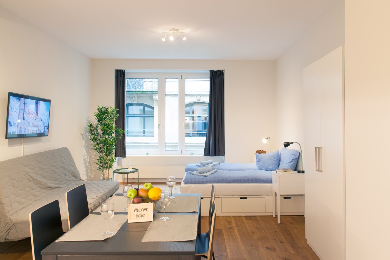 The image shows a studio apartment with a living area, dining area, and sleeping area. The living area has a gray sofa and a TV mounted on the wall. The dining area has a table with chairs and a welcome sign. The sleeping area has a bed with blue bedding