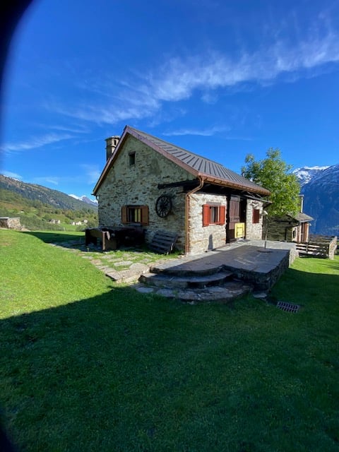 Single story stone house, red shutters, slate roof, porch, wooden wheel, located in a mountainous area with a view of the peaks