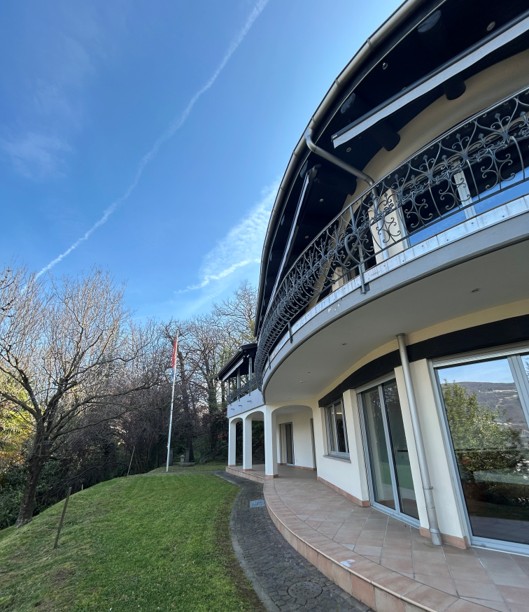 A curved, multi-story building with a balcony or terrace, surrounded by a grassy area and trees.