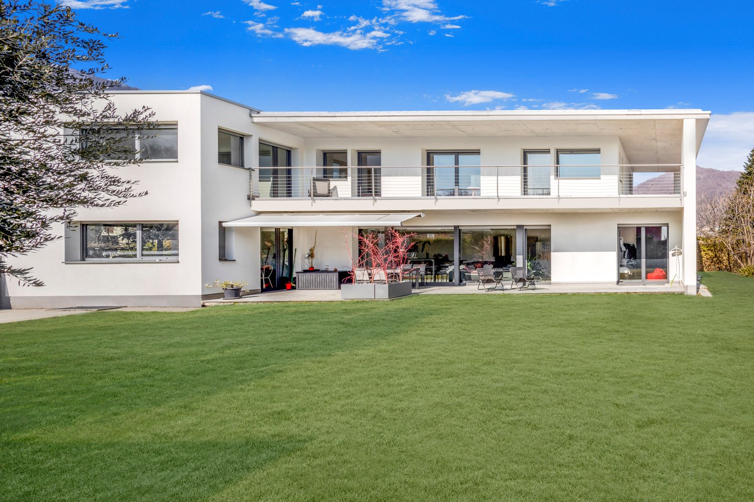 Two-story modern white house with large windows, balcony, and patio area overlooking a well-manicured lawn and garden. The house has a clean, minimalist design with a flat roof and appears to be situated in a scenic, mountainous area with a clear blue sky