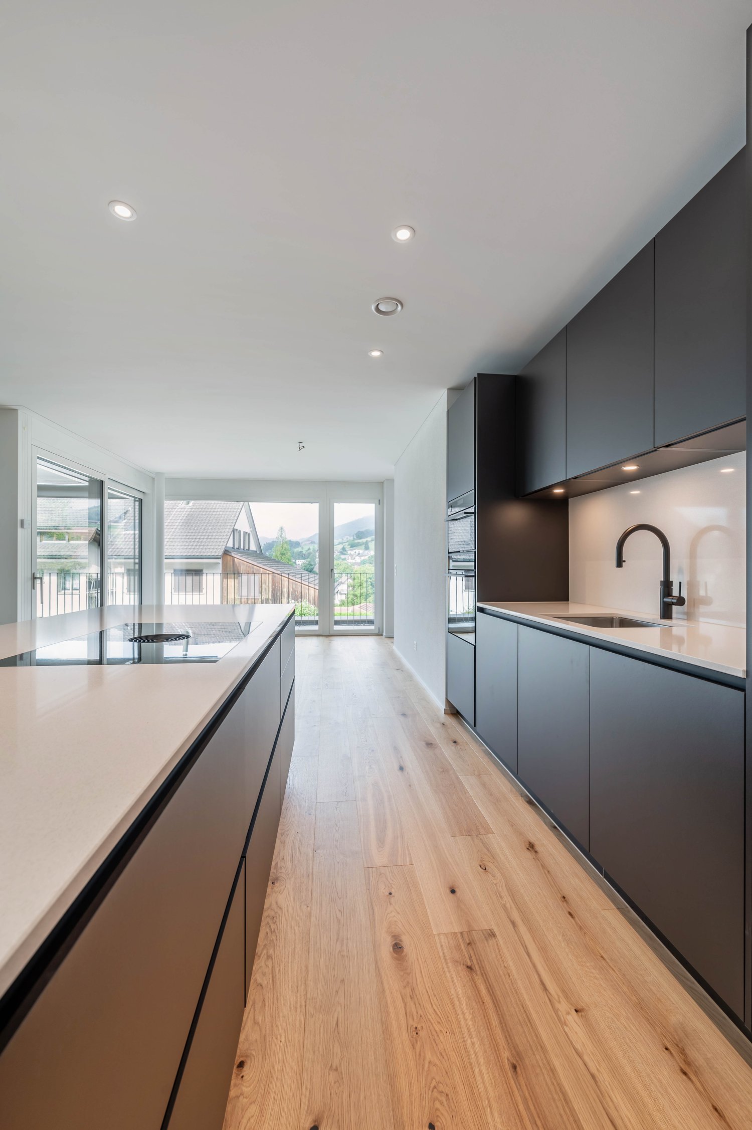 Modern kitchen with oak wood floor, gray cabinets, kitchen island, open windows, balcony, view of the mountains, built-in oven and dishwasher.