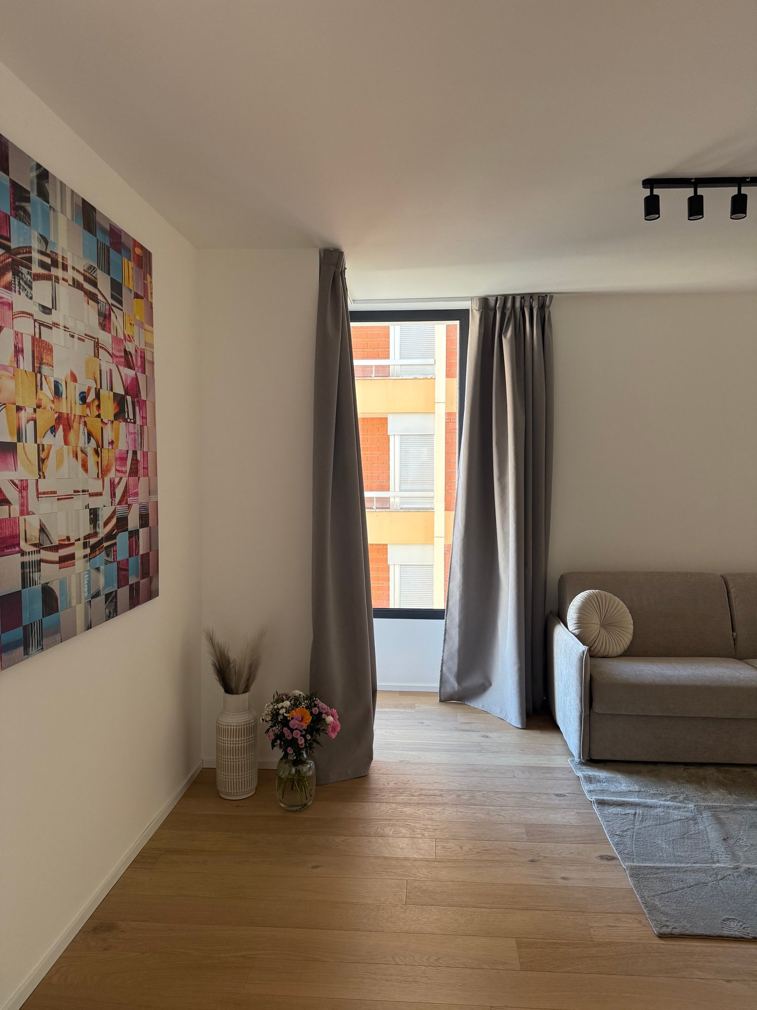 an empty living room with white walls, brown floor, two white vases, one with flowers, gray sofa, rug and large window