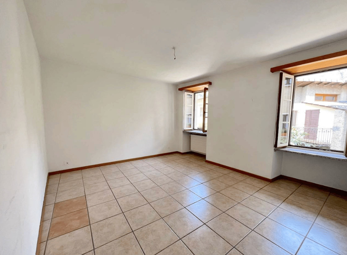 This image shows a spacious room with white walls and a tiled floor. The room has large windows that provide natural light and a view of the outdoors. The room appears to be empty, with no furniture or decor visible.