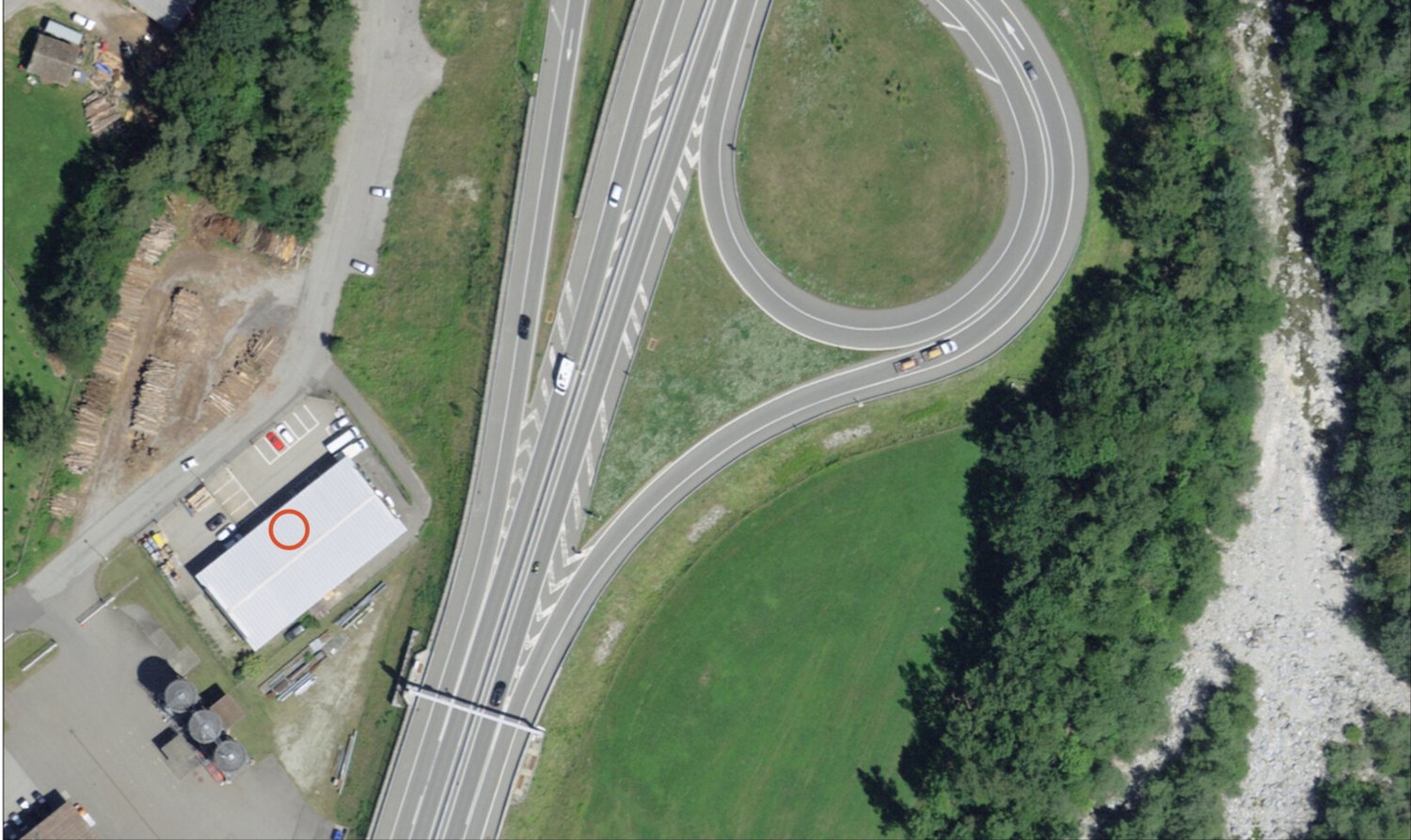 The image shows an aerial view of a highway interchange with multiple lanes and ramps. There is a grassy area in the center of the interchange, surrounded by trees and other vegetation. Several vehicles can be seen on the highway. There appears to be a bu