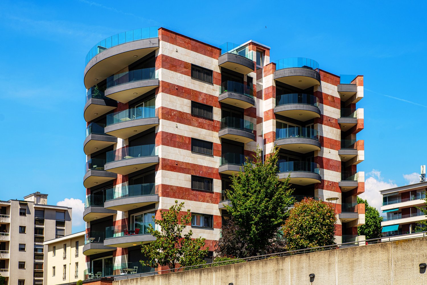 Multi-story apartment building with red brick and glass facade, balconies on each floor, surrounded by trees and other buildings