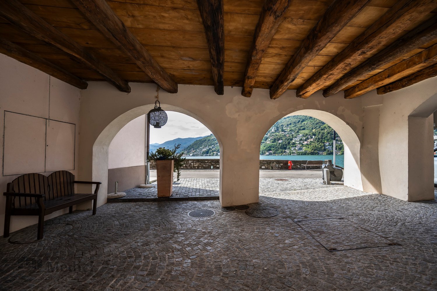 open area with wooden beams ceiling, cobblestone floor, wooden bench, potted plant, two arched openings leading to outside view