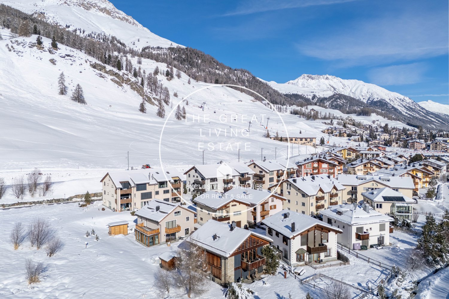 Snowy mountain landscape with a ski resort town nestled in the valley. The town features multi-story apartment buildings and chalets with balconies and parking spaces. The surrounding mountains are covered in snow, creating a picturesque winter scene.