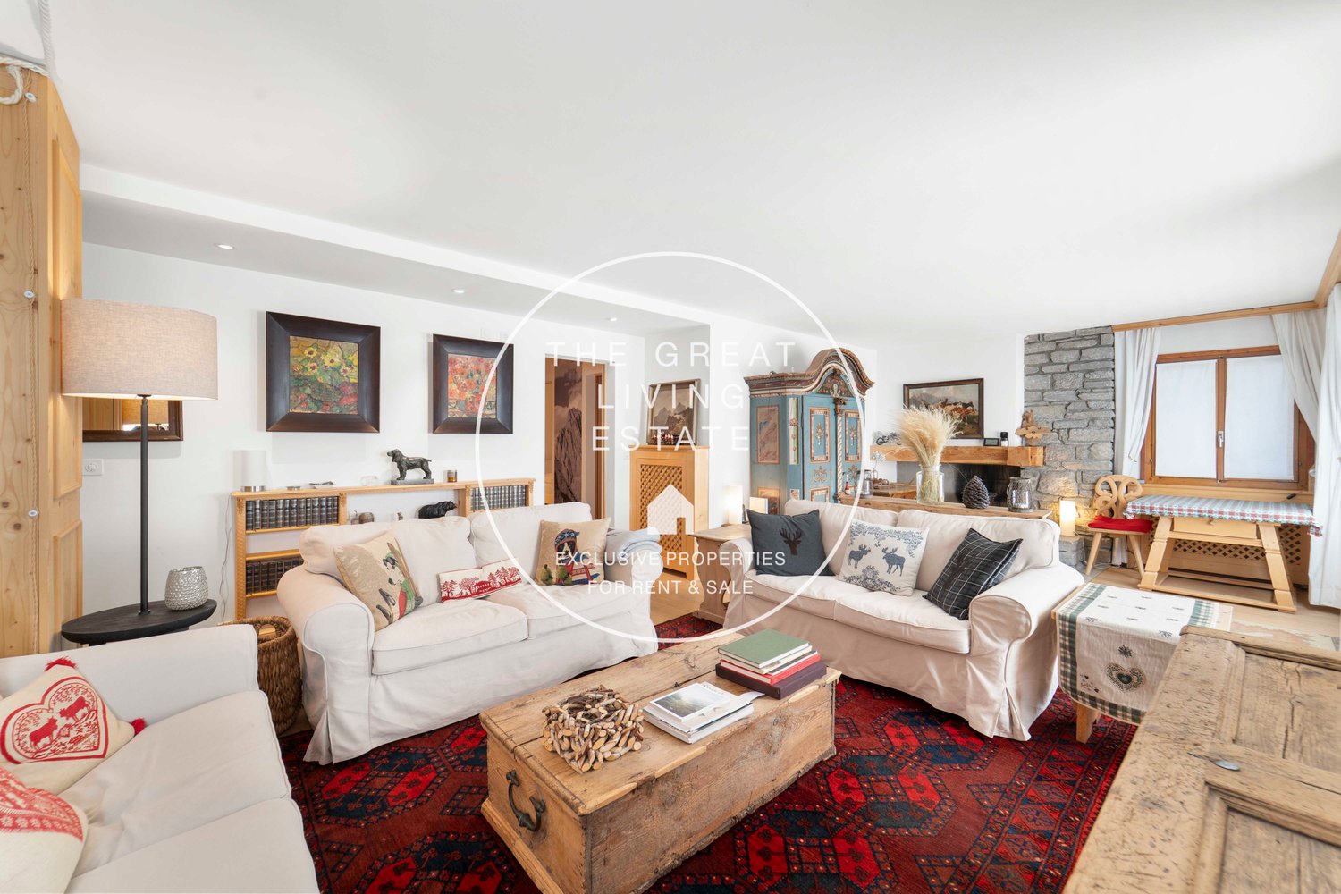 Living room with sofa, coffee table, lamps, bookshelves, decorative pillow, carpet, wooden storage box, decorative objects, stone fireplace, white curtains, paintings