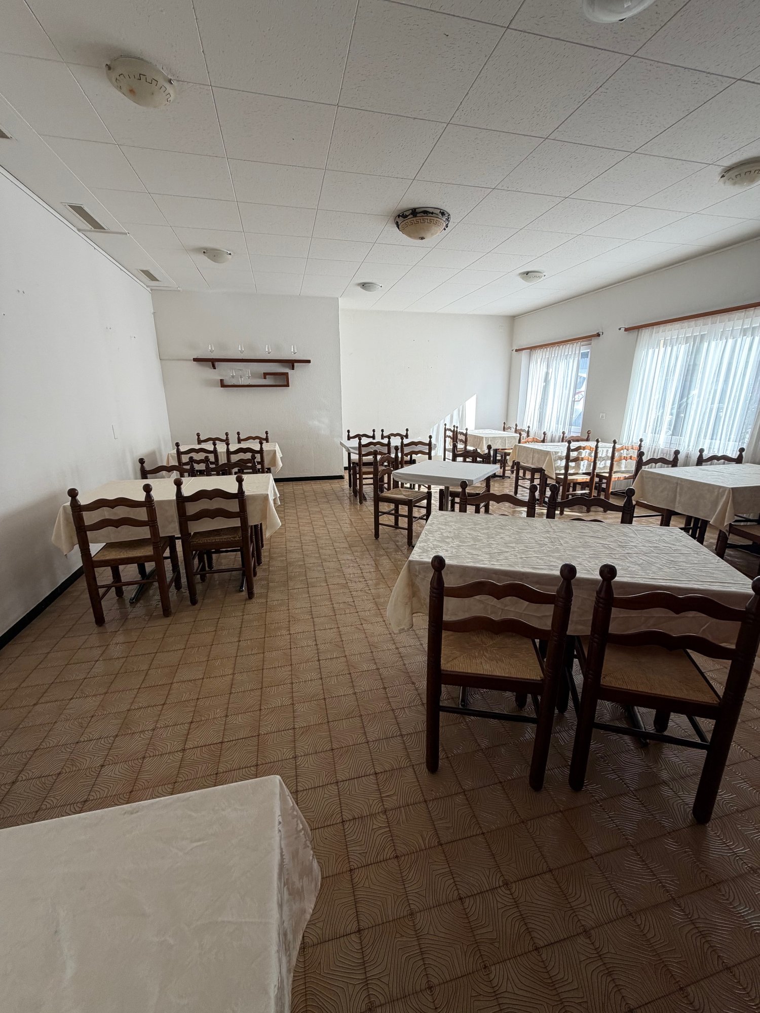 Spacious room, tables and chairs, white walls, tiled floor, overhead lighting, windows with curtains