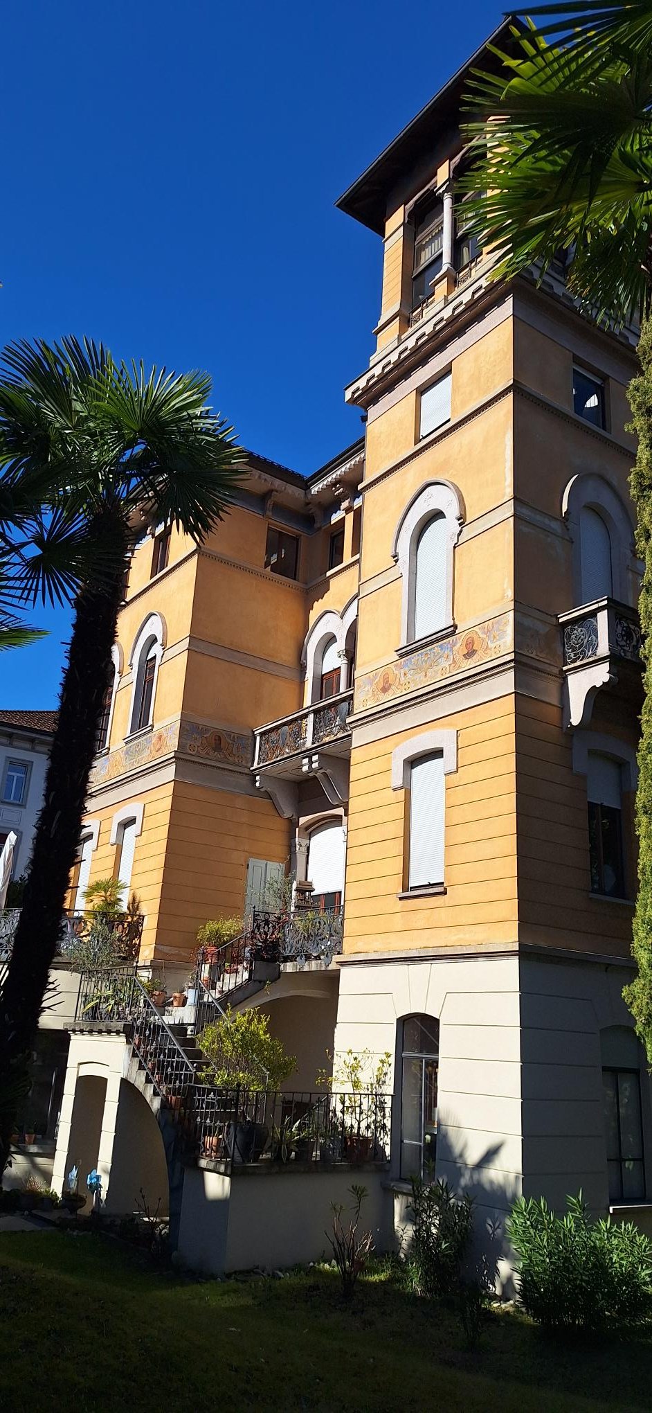 The building has multiple levels, each with balconies, a mix of beige and yellow wall colors, a tower-like section with distinct windows and arches, plants and trees around the area, and a shadow cast on the lower section.