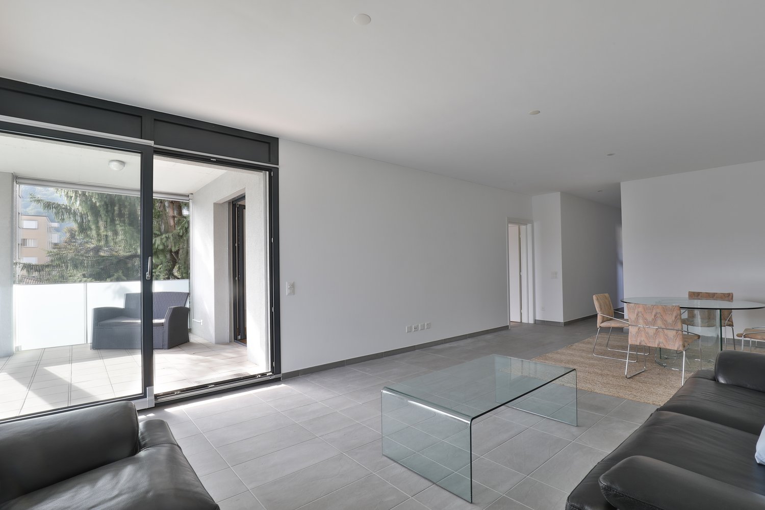 Modern and open living room with sliding glass door leading to balcony, leather couches, coffee table, and dining table with chairs