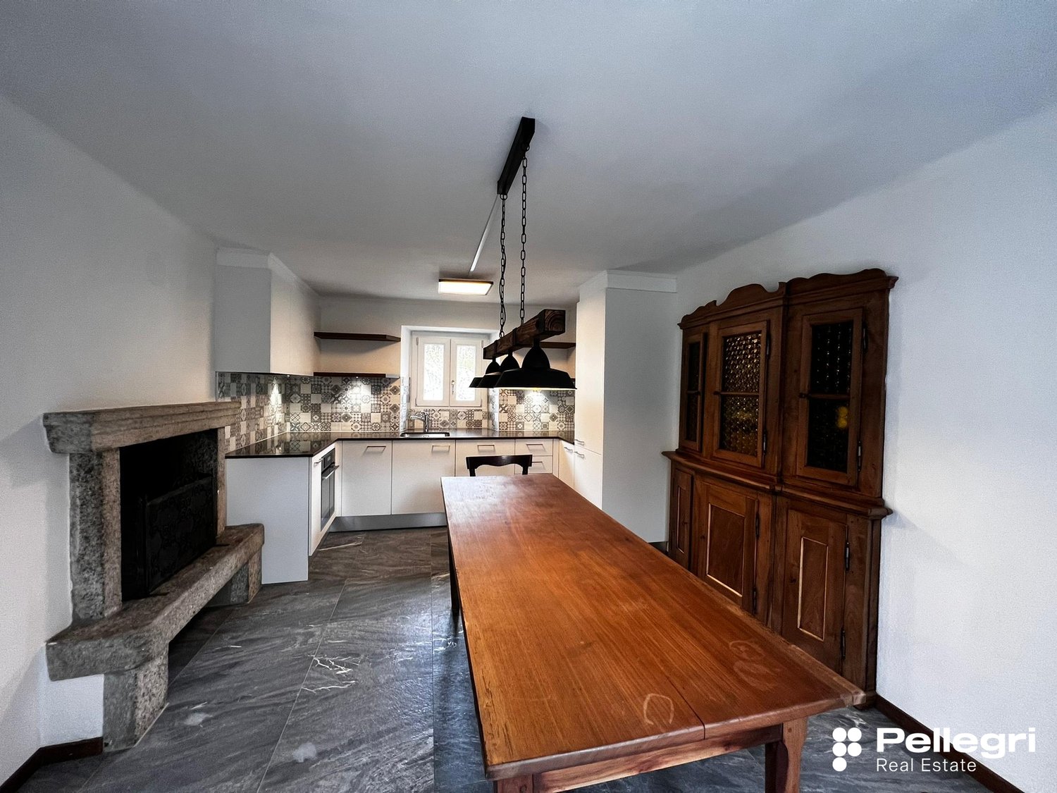 Spacious kitchen with a mix of modern and traditional elements. The kitchen features a large wooden dining table, a built-in fireplace, and a custom tile backsplash. The room also includes a wooden cabinet unit and stainless steel appliances.
