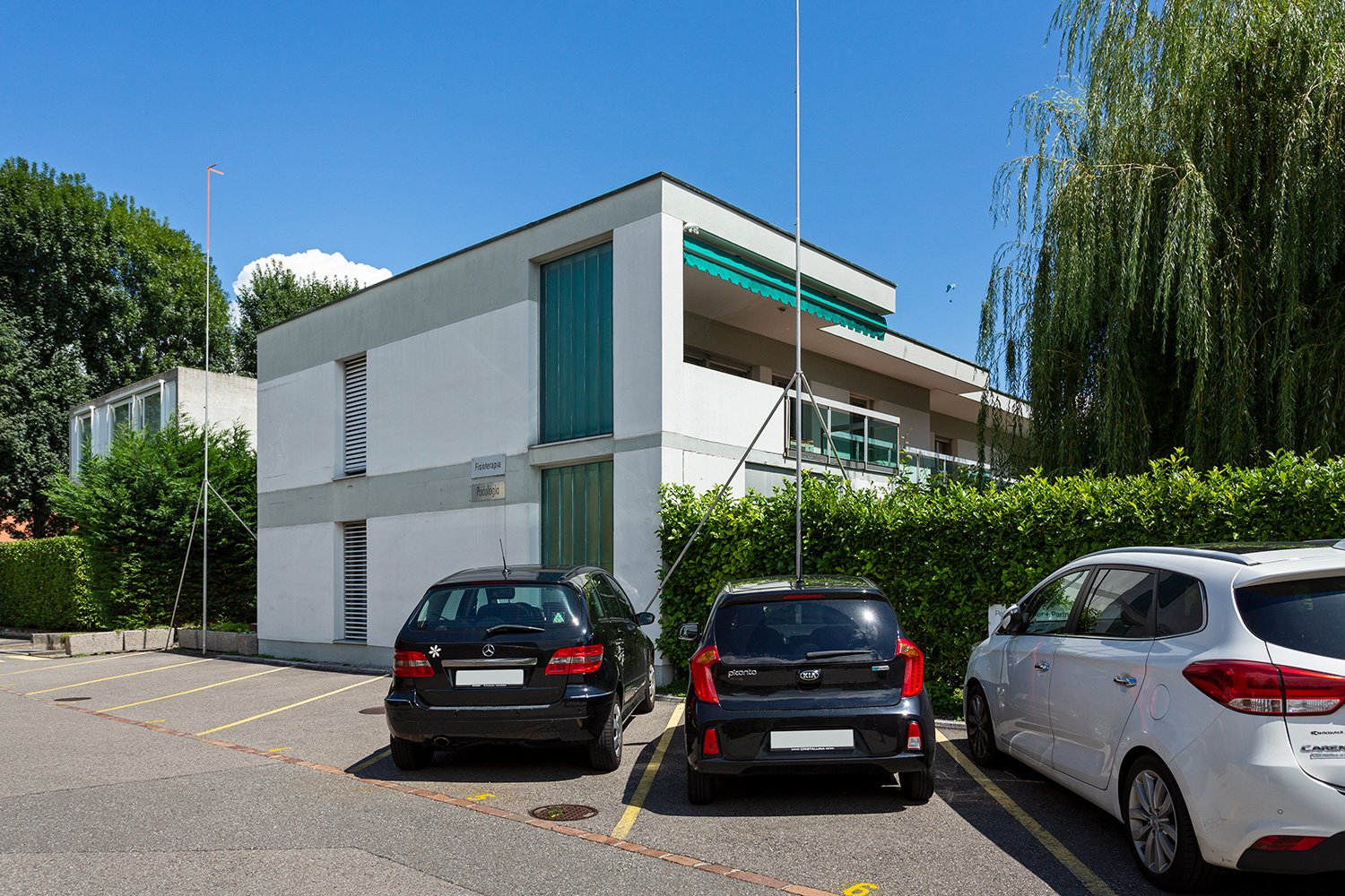 A modern, multi-story building with a gray exterior and green accents. There is a parking lot in front of the building with several cars parked.