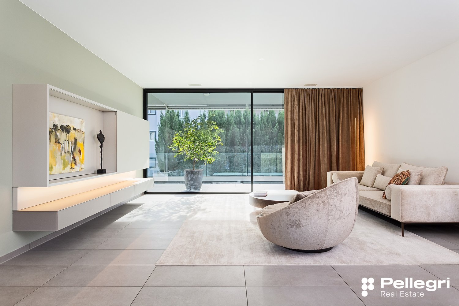 Spacious living room with large windows offering a view of the surrounding greenery. The room features a modern and minimalist design, with a sleek TV console, a comfortable sofa, and a stylish armchair. The curtains add a touch of warmth to the space.