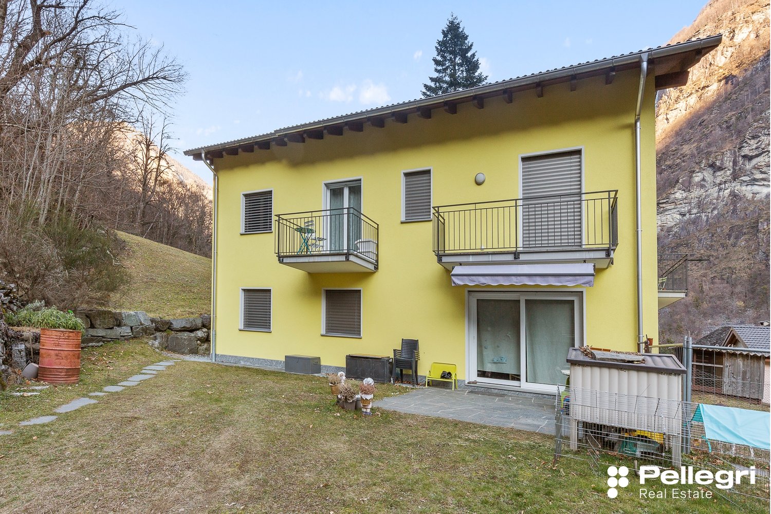 2-story yellow house with balconies, surrounded by trees and a grassy yard, with a rocky cliff in the background