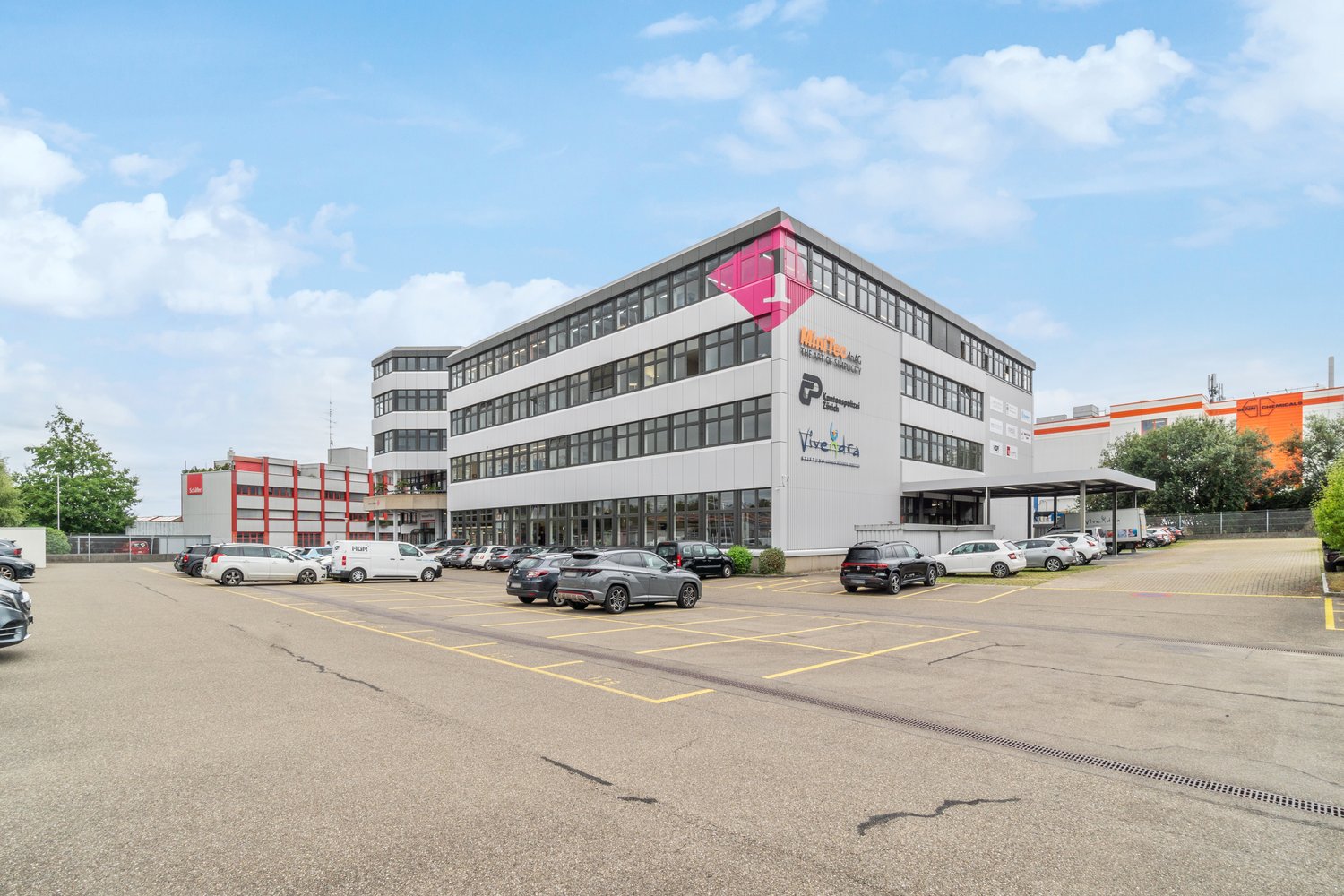 Multi-story office building with glass facade, parking lot in front with several cars parked