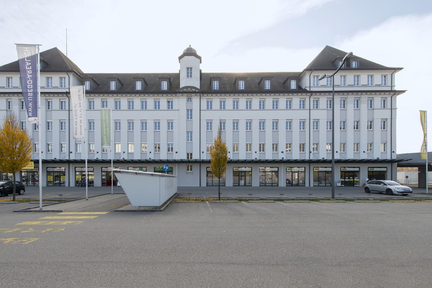 Large multi-story building with a central tower, gray exterior, and several shops or businesses on the ground floor. The building appears to be in an urban setting with a paved area in front of it.