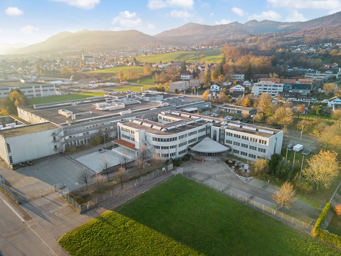 The image shows a large multi-story building complex surrounded by a grassy area and trees. The buildings have a modern, industrial design with flat roofs and large windows. The complex appears to be situated in a hilly, mountainous area with a town visib