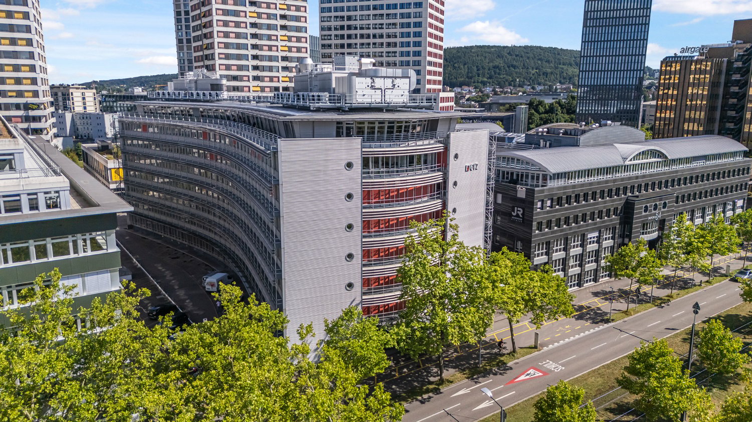 The image shows a cityscape with tall modern buildings, including a curved high-rise building with red accents. There are also other office buildings and skyscrapers visible, along with trees and greenery in the foreground. The scene appears to be a bustl