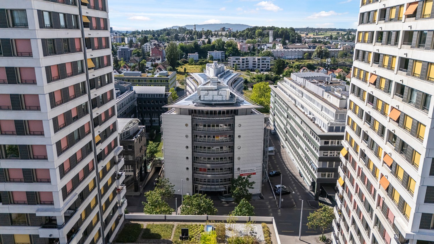 Aerial view of a city skyline with high-rise buildings, residential apartments, and commercial structures. The image shows a mix of modern and older architectural styles, with some buildings having balconies or terraces visible. The cityscape is surrounde