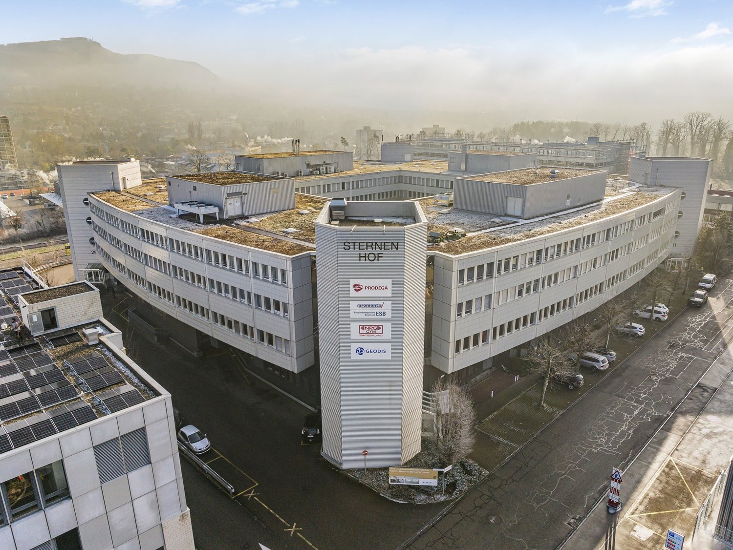 Aerial view of a large multistory office building with a green roof, multiple companies named on the side, surrounded by trees and parked cars