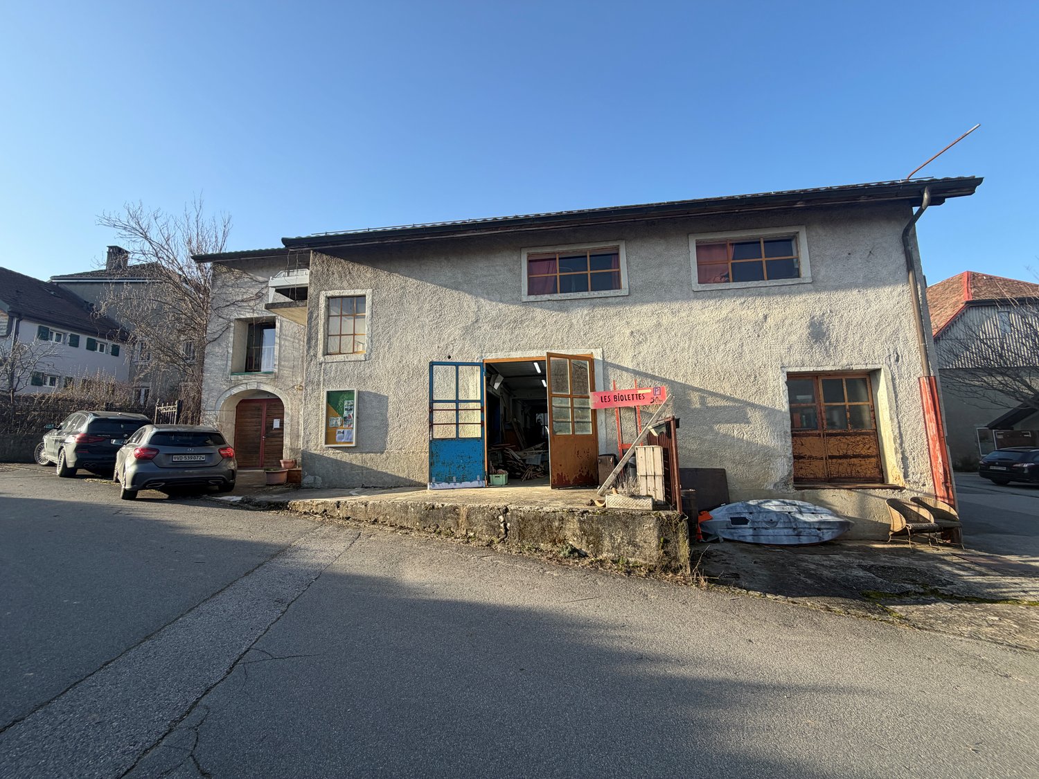 2 story house, concrete exterior, large windows, several cars parked in front, garage entrance, wooden posts, some clutter in the front