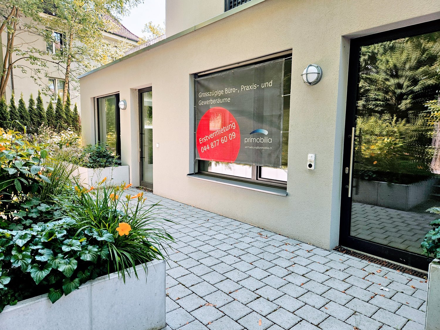 A commercial building with a large window displaying a real estate rental advertisement. The building has a paved walkway and landscaping with plants and flowers in front of it.