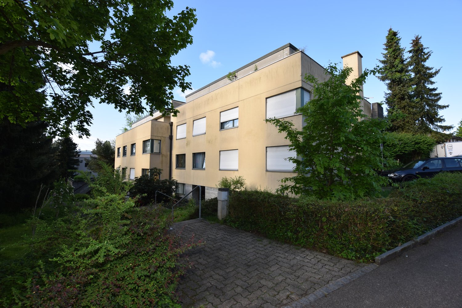 Yellow colored 3-story building with windows with blinds, hedges, trees, and parking.