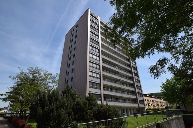 High-rise building with multiple floors and balconies, surrounded by trees and plants, metal fence in front, clear blue sky