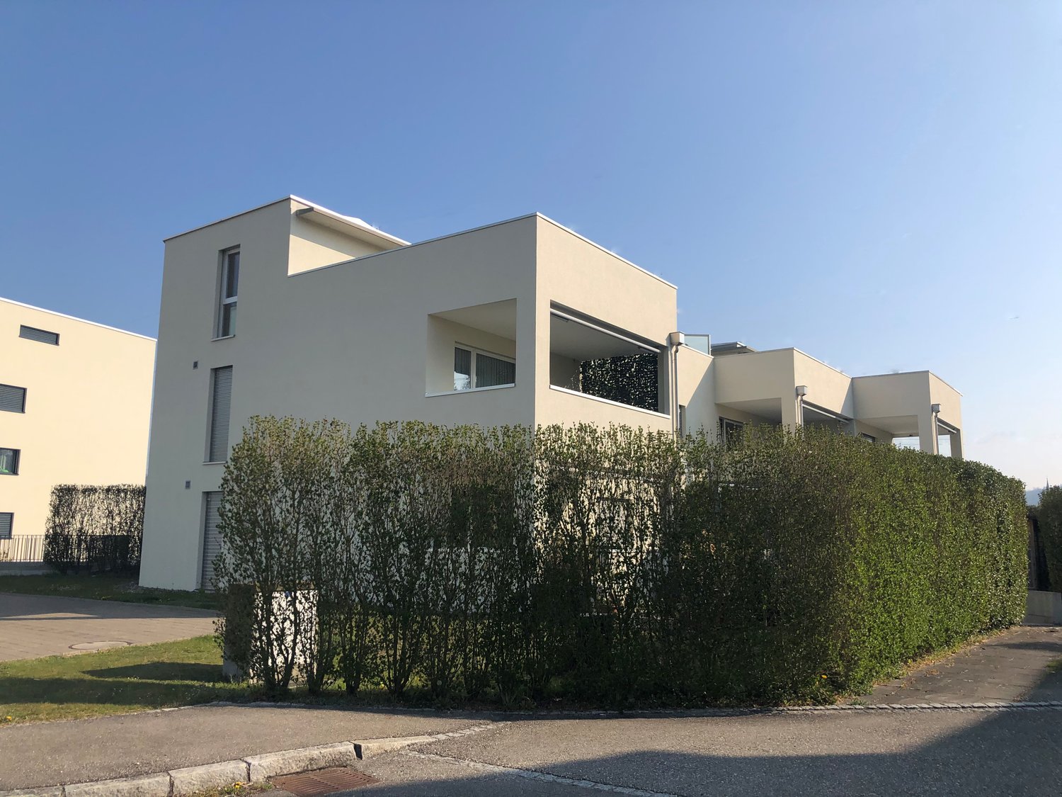 White modern house, 2 stories, large windows, balcony on 2nd floor, 2 doors on ground floor, hedge in front