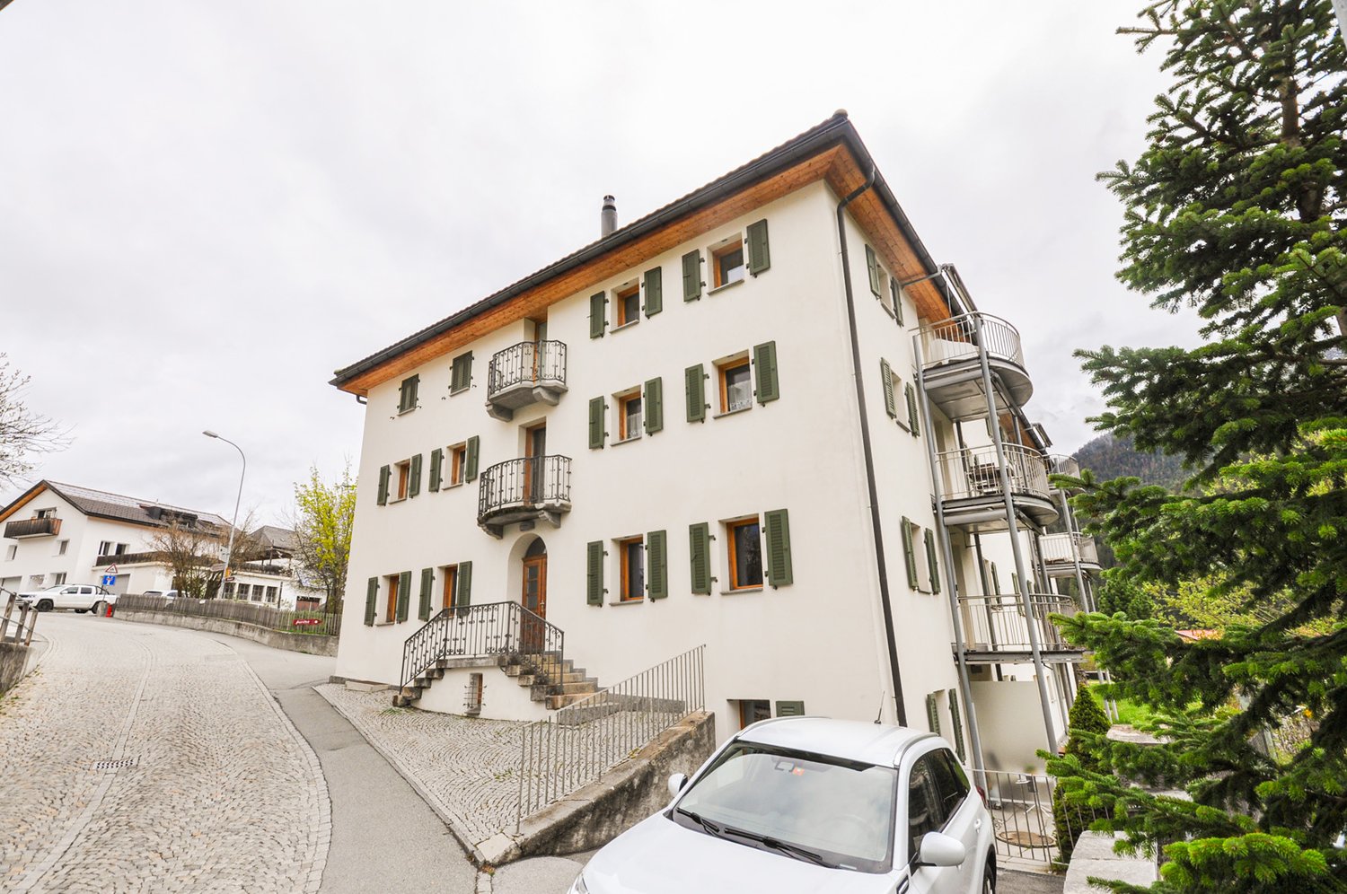 3-story apartment building with white exterior, green shutters, and wooden roof. The building has several balconies and a staircase leading up to the entrance. There is a car parked in front of the building, and the surrounding area is lush with trees.