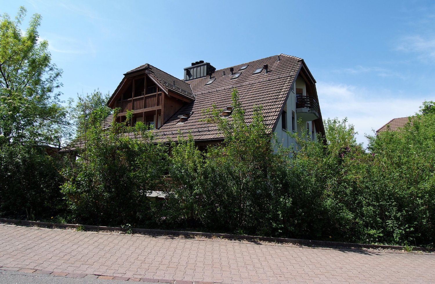 2 story house, brown roof, wooden balcony, brick wall, surrounded by bushes