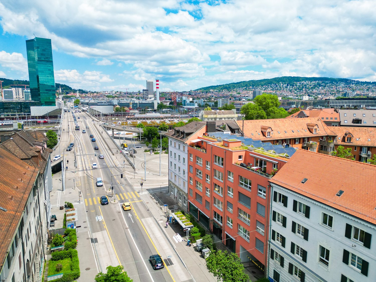 The image shows a panoramic view of a city skyline. There are tall modern buildings, including a green glass skyscraper, as well as older red-roofed buildings. The city is surrounded by hills and mountains in the background. The streets are busy with traf
