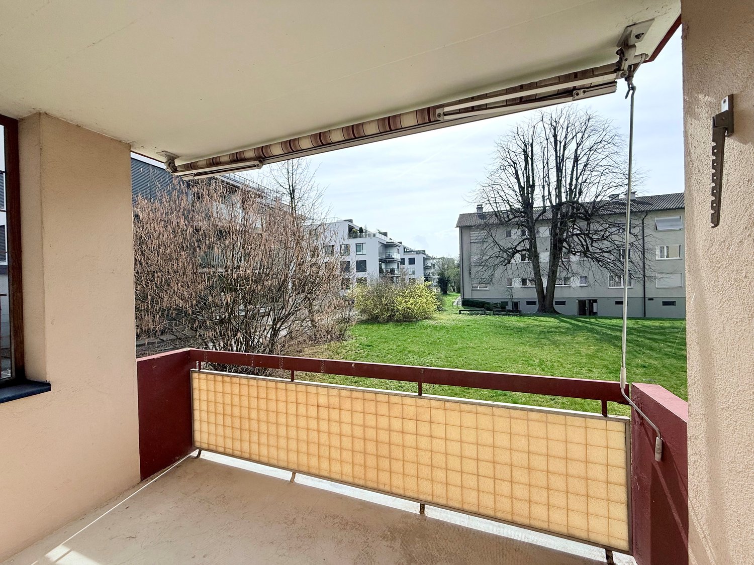 Balcony with a view of a grassy field and trees, red railing, beige shade