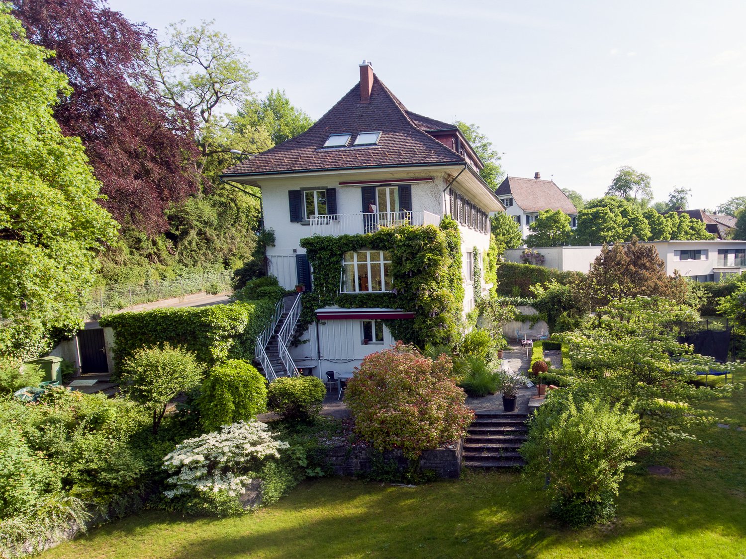 2 story house, white exterior, brown roof, balcony, surrounded by greenery