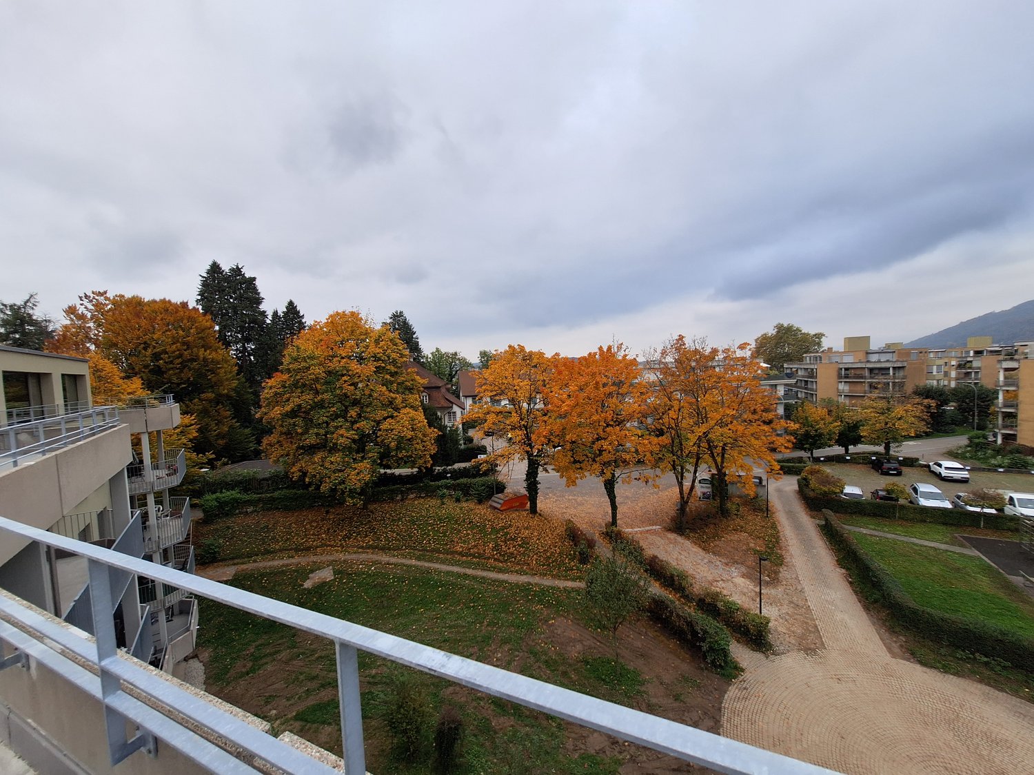 balcony with railings, multiple trees, green grass, concrete pathway, parking area with parked cars, residential buildings, cloudy sky