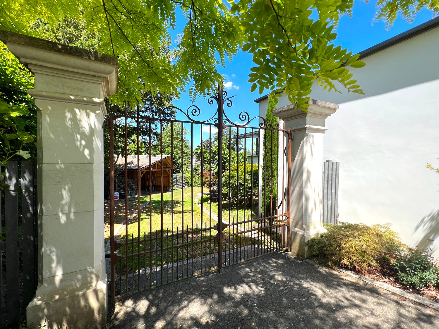Open gate, wide gravel pathway, garden with various plants, trees, and a shed in the background