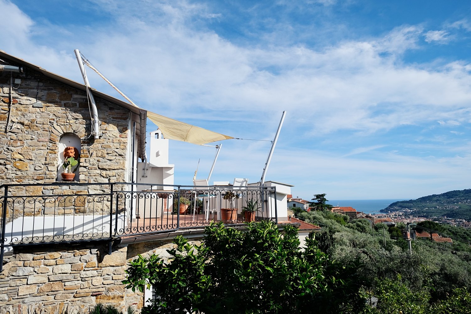 Stone wall building, terrace with awning, potted plants, outdoor table and chairs, scenic view of the ocean and hills