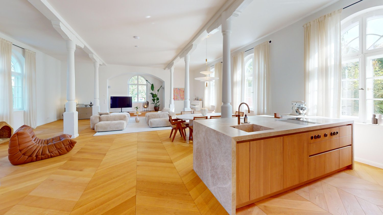 Open-plan living room with a modern kitchen, wooden parquet flooring, central ceiling columns, large windows, dining area, living area with sofas and armchair, television