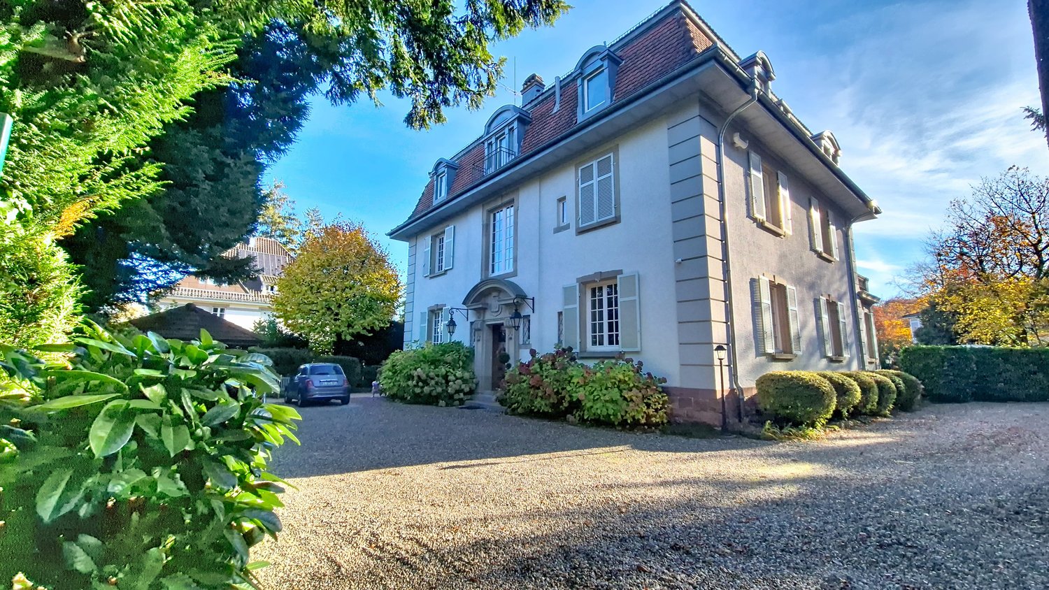 3 story house, white exterior, raised ground floor, gravel driveway, entrance lamps, car parked, garden with shrubs, trees