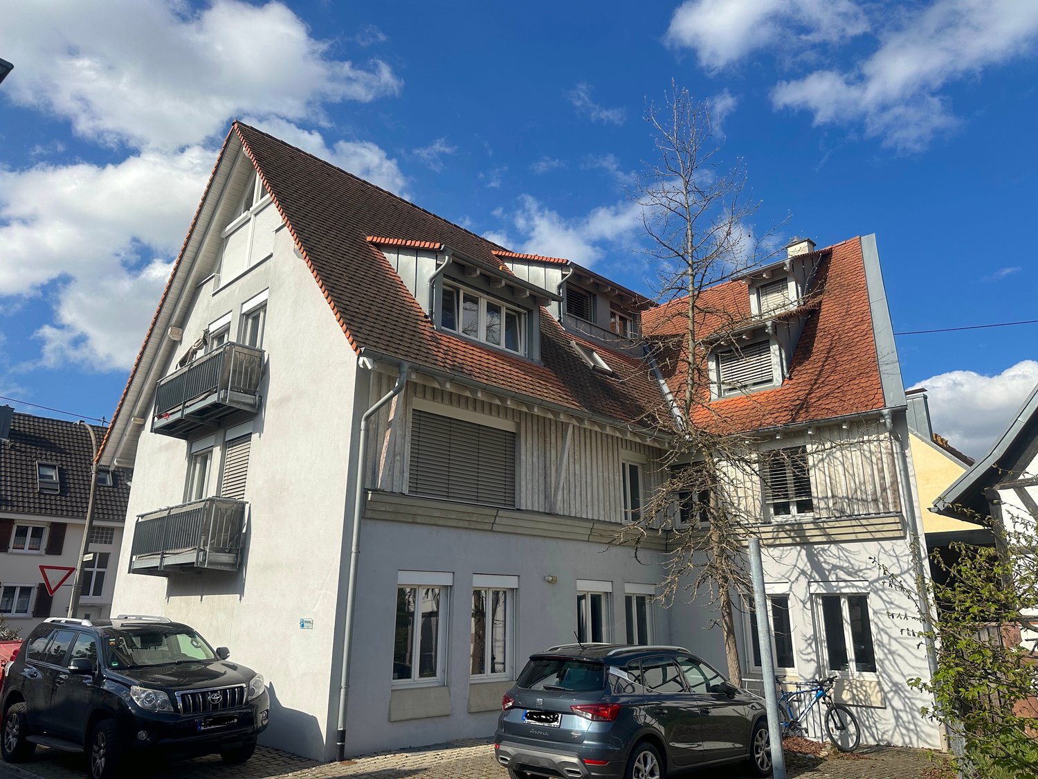 3 story house, white walls, brown roof, balconies, 2 cars parked in front