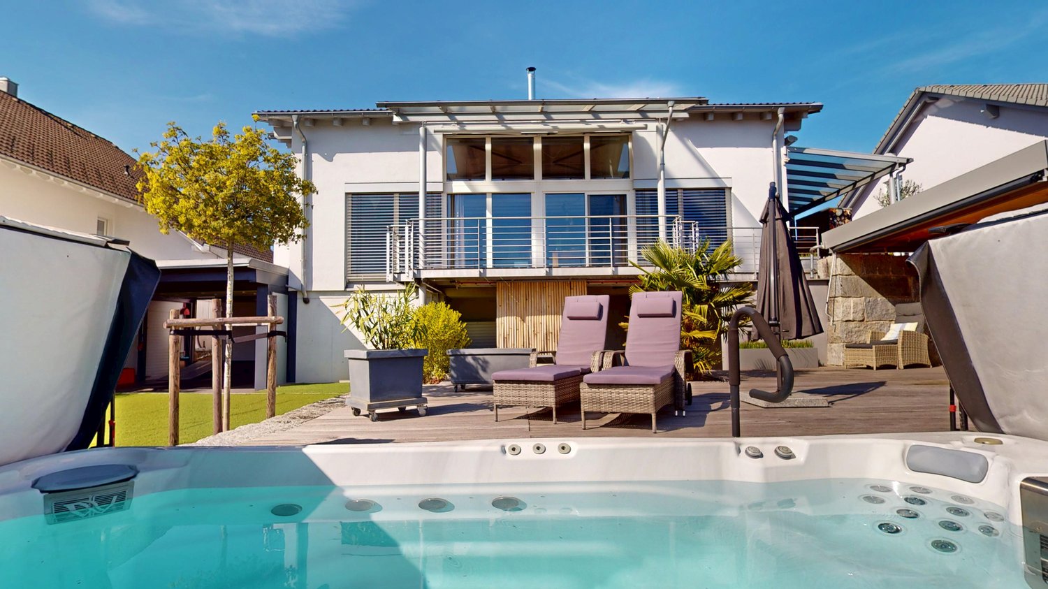hot tub, lounge chairs, umbrella, plants, wooden deck, house in background