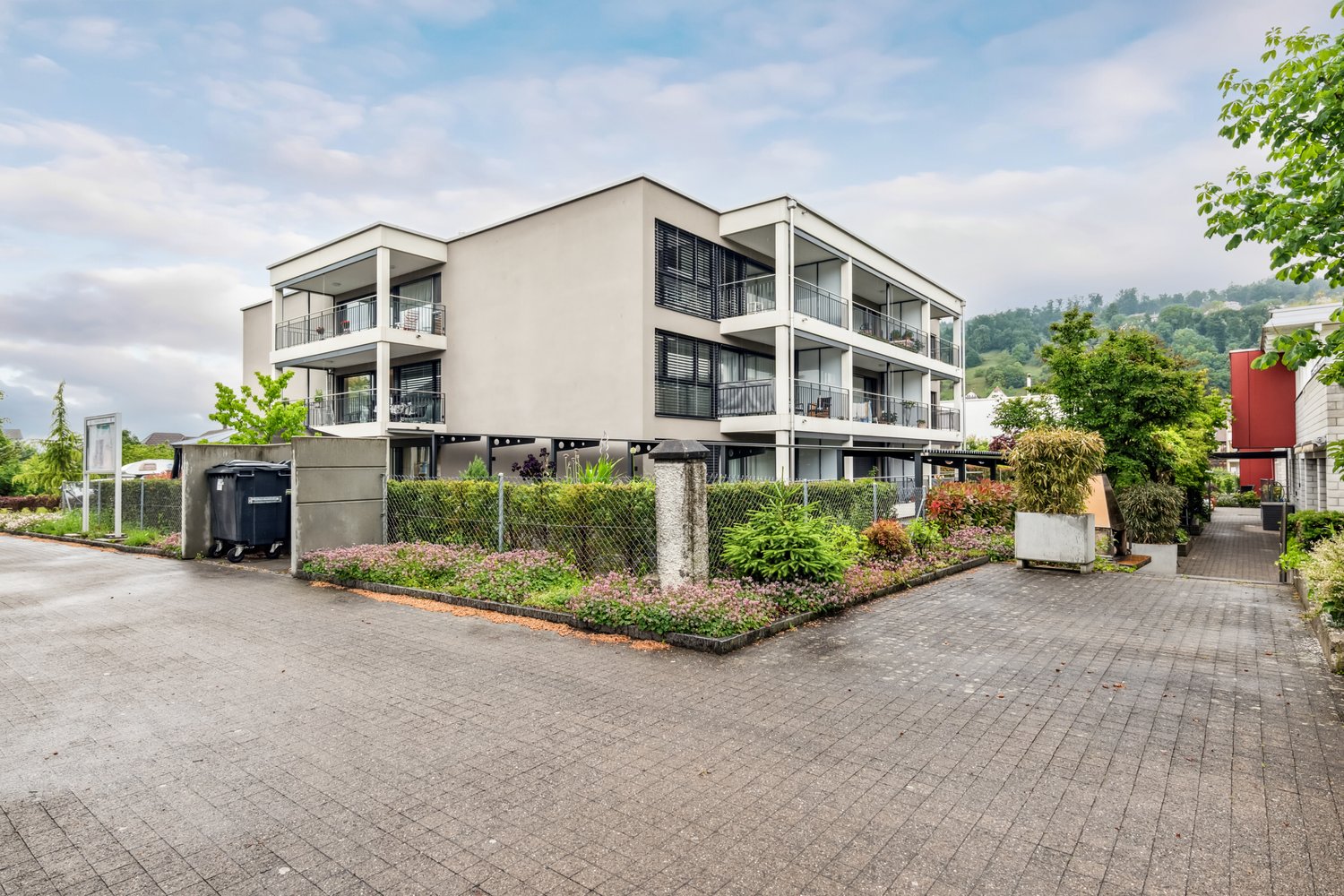 Modern apartment building with balconies, brick walls, paved driveway, greenery