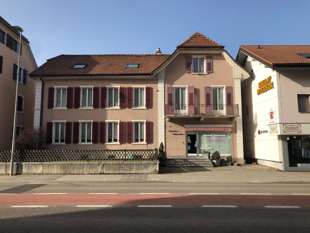 A multi-story building with a red tile roof, pink exterior walls, and white window frames. The building has a balcony on the upper floors and a storefront on the ground level.