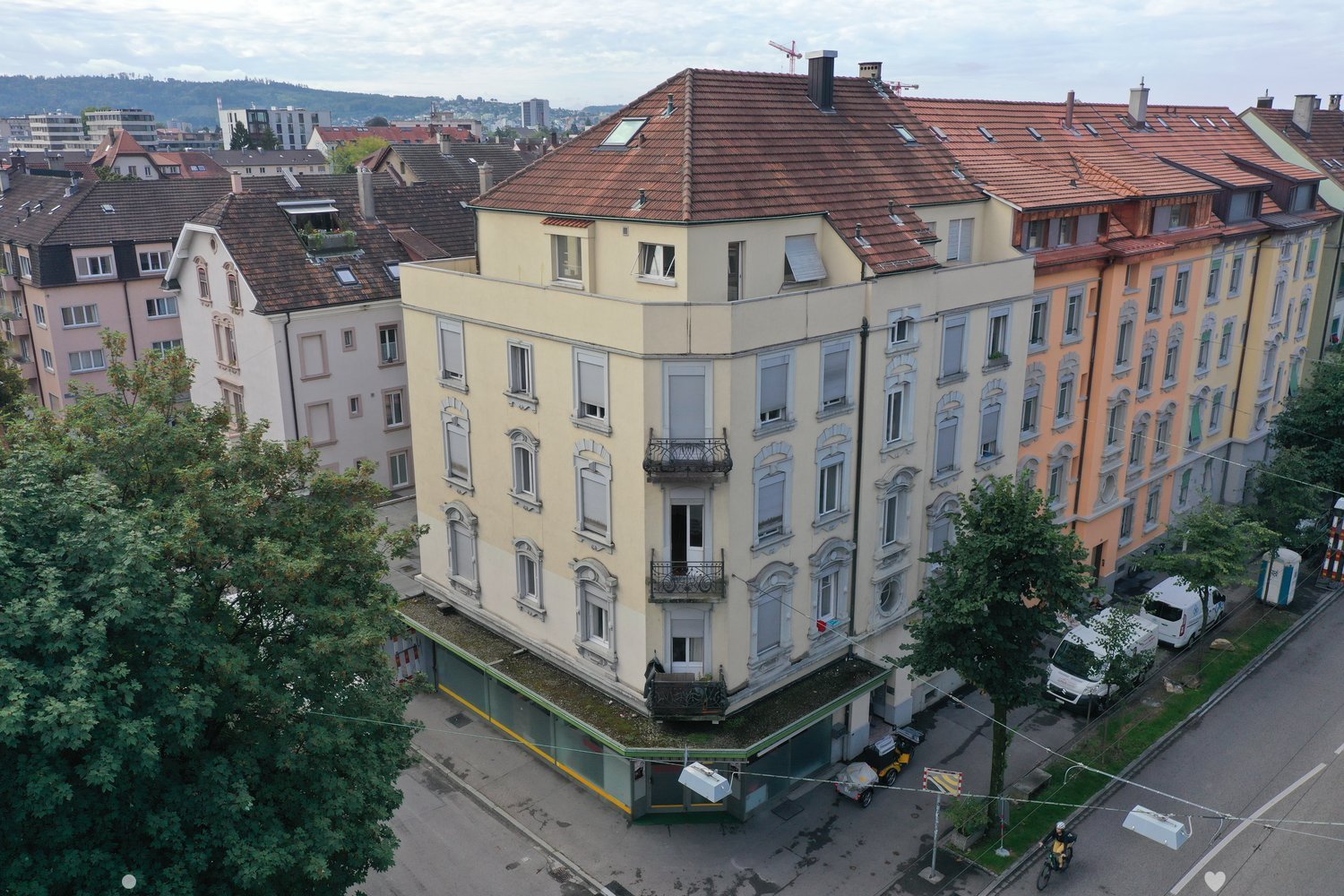 Aerial view of a multi-story building with brown roofs and multiple windows. The building is surrounded by other buildings and trees. There are vehicles parked on the side of the road.