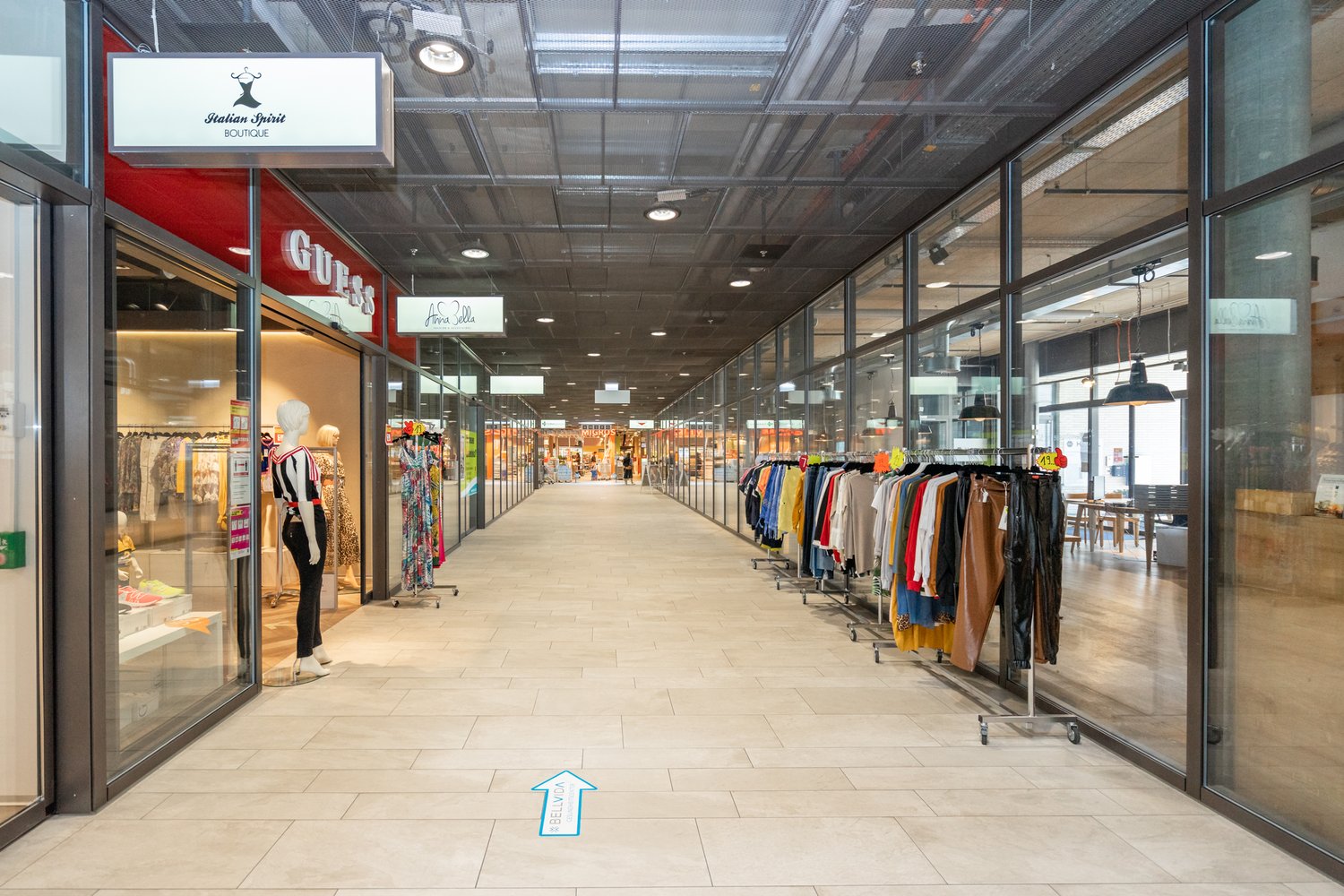 Shopping mall with glass walls, tiled floor, clothing stores, mannequins, clothing racks, and a blue arrow on the floor.