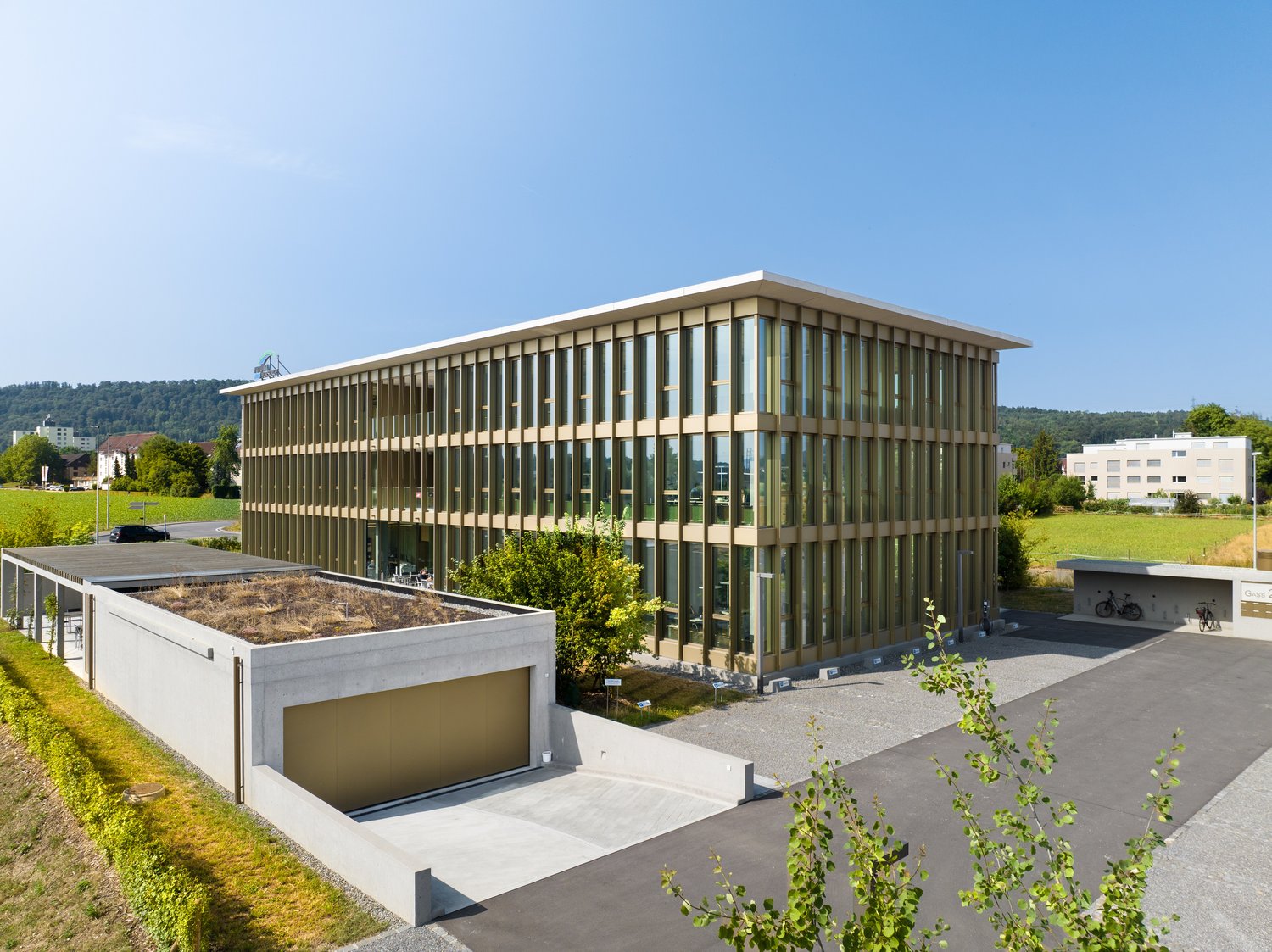 A modern, multi-story office building with a glass facade and a garage on the ground level. The building is surrounded by greenery and has a paved outdoor area with bicycle racks.