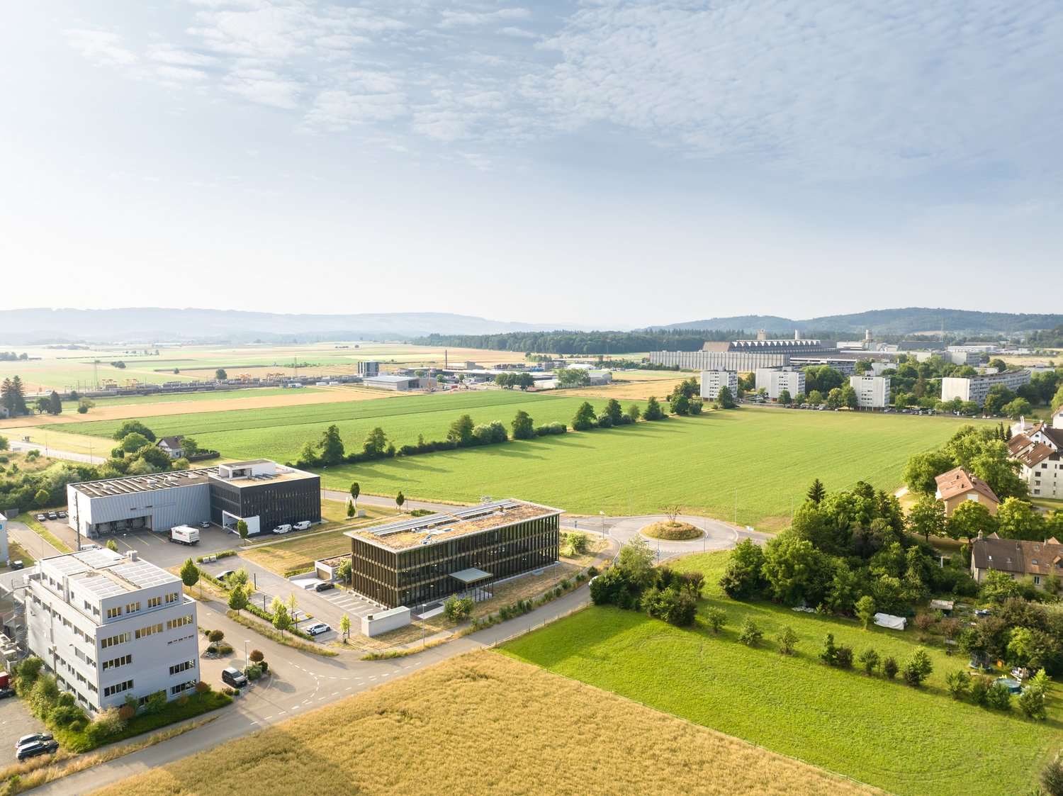 The image shows an aerial view of a suburban area with a mix of residential, commercial, and industrial buildings. The landscape features lush green fields, forests, and rolling hills in the background. There are several large office or industrial buildin