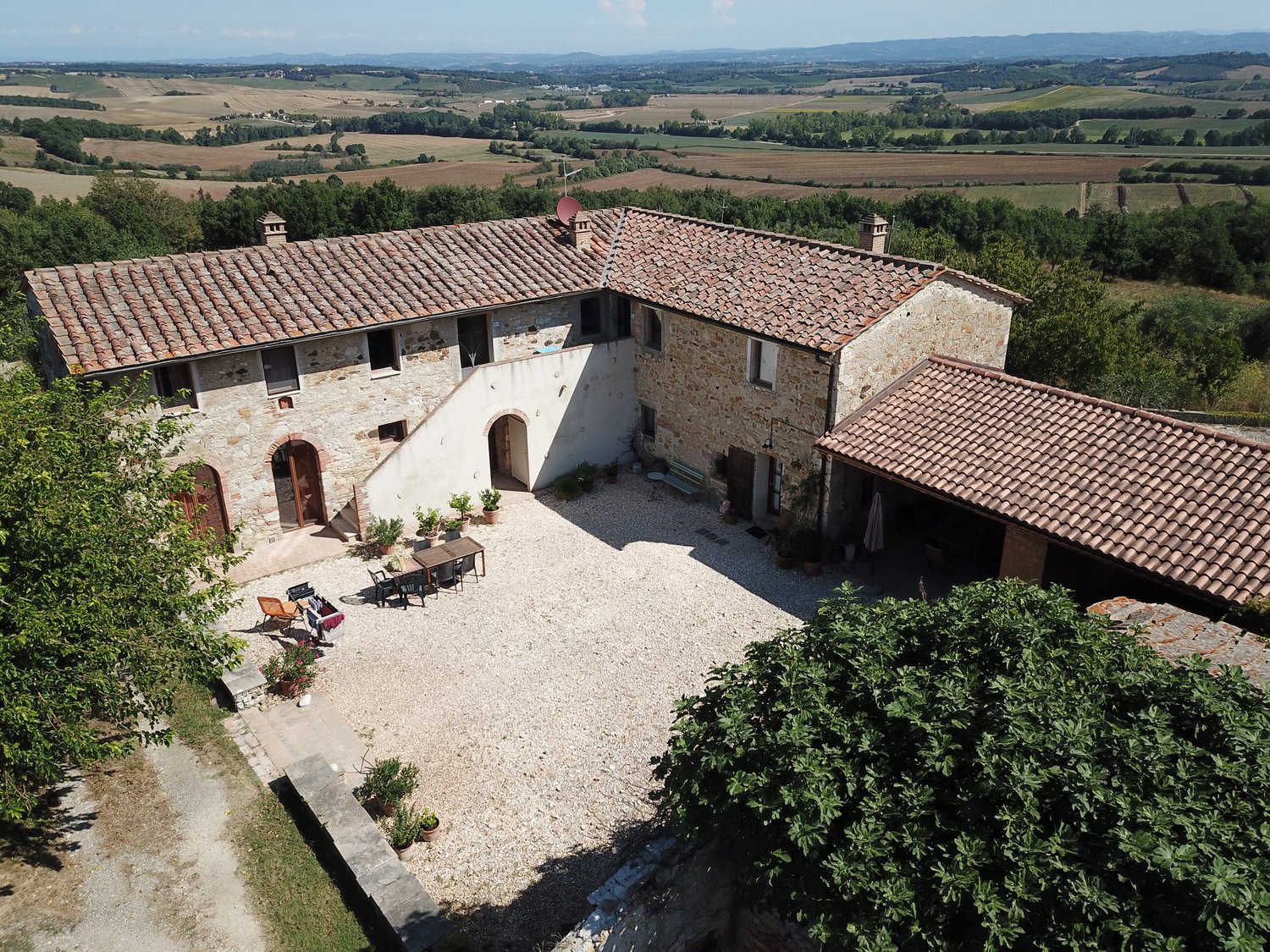 2-story stone farmhouse with red tile roof, courtyard with paved ground, outdoor seating area, surrounded by lush greenery and scenic countryside landscape