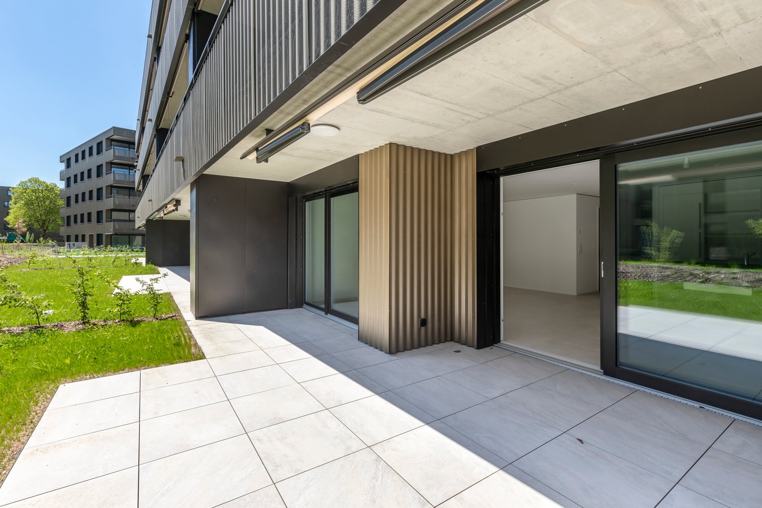 Modern multi-story building with gray metal siding, large windows, and a covered outdoor terrace area with sliding glass doors leading to a grassy lawn